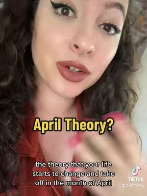 A close-up shot of a woman with long, wavy dark hair. She is wearing a red shirt and dark eyeliner, and her lips are painted with a natural color. A banner in yellow with a black outline and the words "April Theory?" is visible on the image, with the text "the theory that your life starts to change and take off in the month of April" below it.