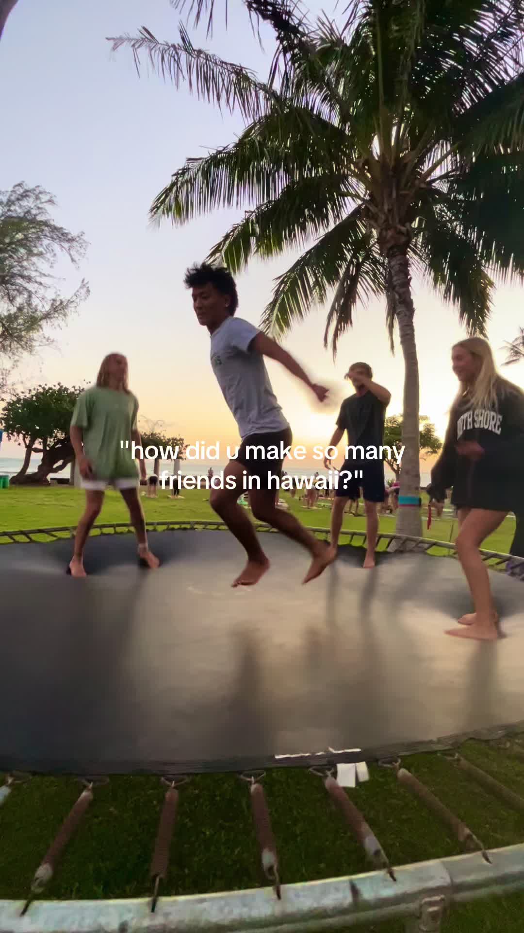 A group of young people jumping on a trampoline outdoors. The scene is set at sunset with palm trees and a grassy area in the background. A text overlay asks how the creator made so many friends in Hawaii.