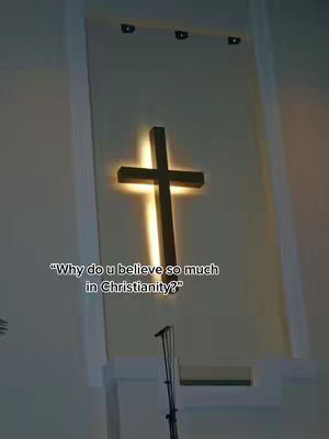 A dimly lit image shows a church interior with a cross illuminated on the wall. The cross is a dark silhouette against a bright, glowing background. Below the cross, there is a microphone stand, and a text overlay asks "Why do u believe so much in Christianity?"