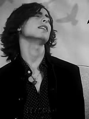 A grayscale image showing a young man with long hair, eyes closed and head tilted up. He wears a patterned shirt under a dark jacket and a heart-shaped pendant necklace. The background is blurred with a suggestion of birds flying.