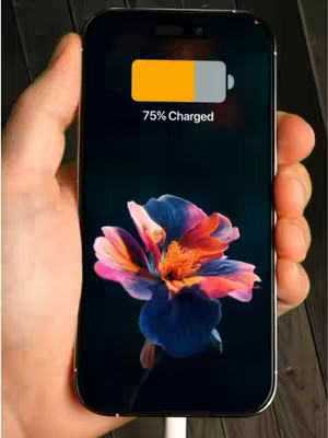 A hand holding a black iPhone displays a colorful floral live wallpaper. The phone's battery shows 75% charged. The background is textured wood, placing the phone in an indoor setting.
