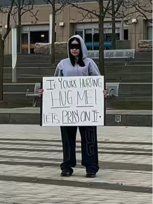 The image features a person standing in a city location, holding a sign. The person is wearing a grey hoodie, dark pants, and sunglasses. The sign reads, "If You're Hurting, Hug me! Let's Pray on it." The background includes urban architecture, greenery, and a paved area. The lighting appears to be daytime, and the overall tone is one of offering support and community.