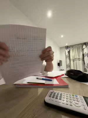 The image shows a person's arm holding up a sheet of paper with handwriting or print, partially obscuring their face. The foreground features a desk with a notebook, pen, and calculator on it in a dimly lit room.