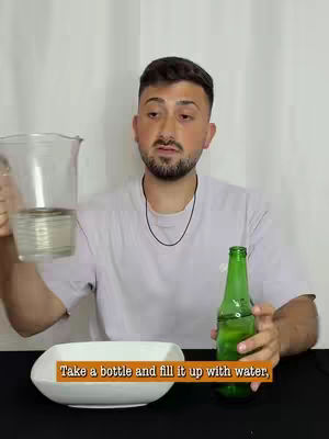 A medium shot of a man standing in front of a white backdrop. He is holding a clear glass beaker in one hand, partially filled with a liquid. A green beer bottle is in his other hand, a white dish sits in front of him. Text overlayed on the image is 'Take a bottle and fill it up with water'.