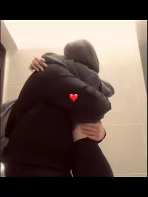 A couple embracing each other in a close hug. The image is somewhat dimly lit, with the subjects appearing in dark attire set against a plain background.