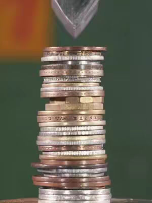 The image shows a stack of coins of different sizes and colors, with a silver object poised above them. The background is a mix of green, orange, and yellow.
