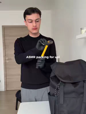 A young man holding a hammer over a backpack, preparing to pack. The background is a simple room with a door and a white shelf. Text overlay reads 'ASMR packing for uni'.
