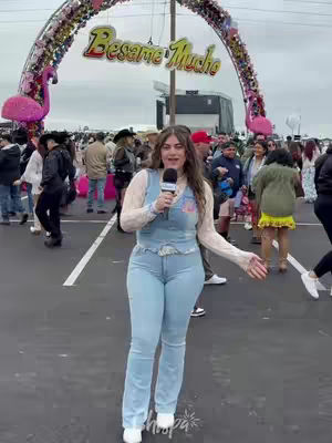 A woman stands in the center of a bustling, outdoor concert or event area. She's holding a microphone and gesturing with her arm, with a sign over her shoulder.