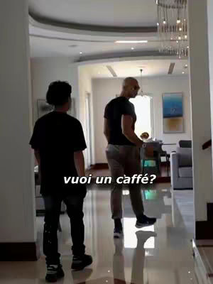 Two men are walking in a luxury house. The man in the front is wearing a black t-shirt and dark colored pants, and the other man is wearing a black t-shirt, shades, and light colored pants. The floor is glossed concrete and there is a chandelier in view.