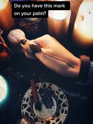 A hand, adorned with black nail polish and a decorative wristlet, holds a crystal ball while resting on a surface surrounded by candles, a decorative plate, and other occult-themed items.
