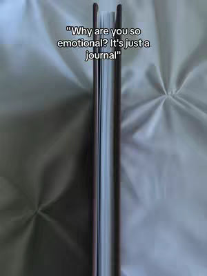The image shows a journal held upright, with the spine facing the camera. The journal is closed, and the edges of the pages can be seen. The background is a blurry, light-colored fabric, possibly a bedsheet. Text overlayed on the image says, "Why are you so emotional? It's just a journal."