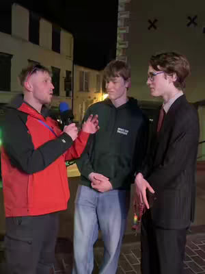 A nighttime street scene with three young men. A man on the left, holding a microphone, appears to be interviewing the other two. The background features buildings with lit windows.