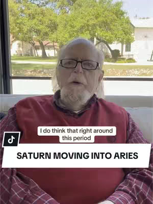 An older man with glasses and a gray beard is seated on a light-colored couch. His face is centered, with a red shirt peeking from under a checked shirt. Behind him, a natural outdoor scene is visible with green trees and a building. Overlaying the image, a white banner reads 'SATURN MOVING INTO ARIES', with a smaller text box above it saying 'I do think that right around this period'.