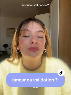 A young woman with fair skin and long, light brown hair, wearing a light yellow sweater, is centered in the frame. She has a warm expression with slightly parted lips, and her eyes are visible, as if looking directly at the viewer. She's wearing small silver hoop earrings. Overlayed on the bottom is a light purple rounded rectangle containing the text "amour ou validation ?" in white; a small music note symbol is in the top-right corner of the rectangle and the word 'aime' is at the bottom.