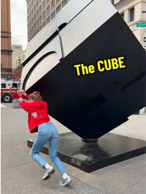 A woman in a red jacket and blue jeans appears to be pushing against a large, black and white sculpture in a city environment. Text overlayed in yellow reads 'The CUBE'. In the background, there are buildings and a fire truck.