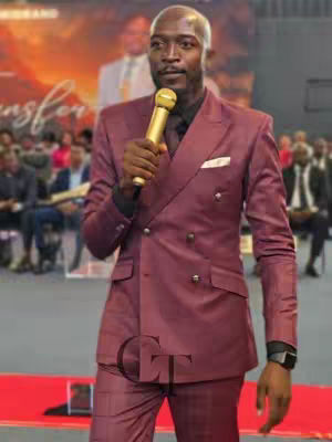 A man in a burgundy suit is holding a golden microphone against a blurred background. He is standing on what appears to be a stage with a red carpet, and there is a blurred audience behind him, with a glowing logo on the background.