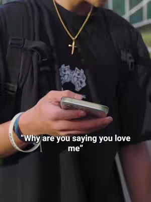 The frame captures a person from the chest up, holding a phone in their right hand. Their attire includes a black shirt, a black vest-like accessory with pockets, and a gold chain with a cross pendant. A white and blue bracelet adorns their wrist. The background is slightly blurred, featuring hints of green. Overlay text is near their hand.