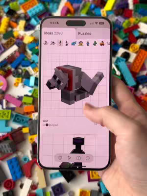 A smartphone is held up showing a 3D model of a wolf from Minecraft, composed of pixelated blocks, against a pink background. The phone is surrounded by a scattered collection of colorful LEGO bricks, and a hand is visible holding the phone. The app's interface includes options and a progress bar at the bottom, and a 'Puzzles' tab is displayed, suggesting it's a building app based on the Minecraft game.