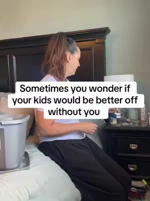 A woman with brown hair pulled back in a ponytail sits on a bed, slightly turned away from the camera. She's wearing a white t-shirt and dark pants. In the background is a large black headboard and a nightstand with items on it. The overlay text reads, "Sometimes you wonder if your kids would be better off without you."