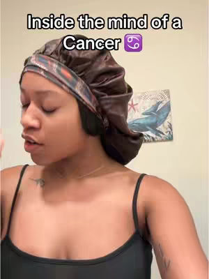A person in a brown satin bonnet is shown from the chest up, in front of a cream-colored wall. The person looks down with a slight expression on their face to the left of the frame. Below the person, dark spaghetti straps are visible. Behind the person, a framed piece of artwork is visible. The artwork depicts a blue whale and a few red stars, and is against a white background. Above the person, superimposed text reads, "Inside the mind of a Cancer".