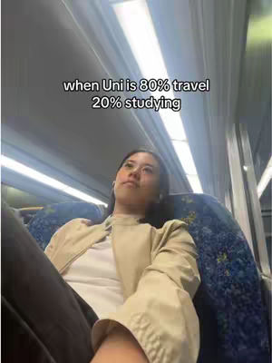 A selfie video taken on a train. A young woman is seated and looking up, seemingly tired. Text overlayed on the image reads: 'when Uni is 80% travel 20% studying.' The background shows the interior of a train, with overhead lighting panels. Seating consists of blue seats. The composition is candid, with the subject centered and the background slightly out of focus.