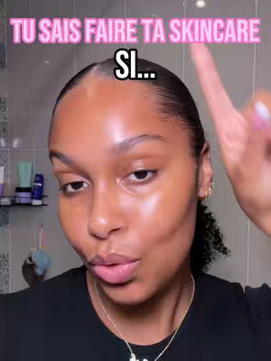 A selfie-style shot featuring a woman looking towards the camera. She has smooth, glowing skin and is wearing a black top with a delicate necklace. Her hair is slicked back, and she appears to be in a bathroom setting, with some toiletries visible in the background. The image is well-lit, and she is pointing her finger, giving gesture.