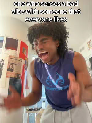 A young man with curly hair, wearing a blue tank top, and light-colored pants is captured in a moment of laughter. His mouth is wide open, and his eyes are crinkled, suggesting genuine amusement. The background includes a cluttered room with posters on the wall.