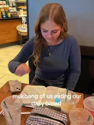 A young woman sits at a table in a Starbucks, looking down at an open Bible. There are drinks and a small purse on the table. The background includes a cafe counter and other tables.