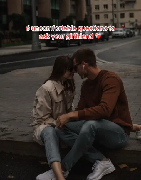 A couple sits close together on a concrete ledge outdoors, their faces touching and eyes closed intimately. They are surrounded by buildings and a street. The text overlay says, "6 uncomfortable questions to ask your girlfriend."