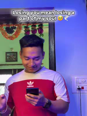 The image captures a young man indoors. He is wearing a white t-shirt with a red panel across the chest, and the Adidas logo is visible. He is holding a phone in his right hand, looking down, and appears to be interacting with the device. The background includes a wall, with some decorative elements like ornaments and a mirror. The person has fair skin and dark hair. The overall lighting seems natural, with no special effects.