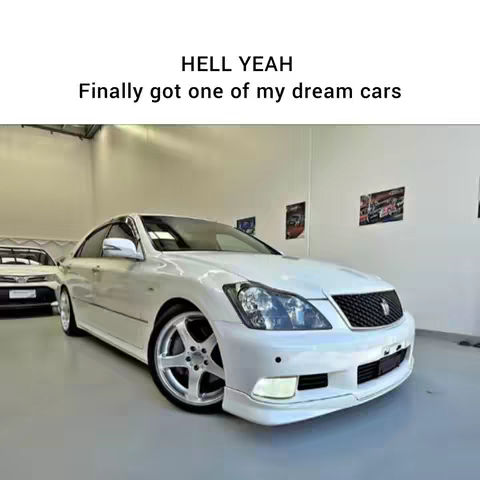 A white Toyota Crown Athlete is parked inside a well-lit showroom. The car takes up most of the frame, with its front angled toward the viewer. It has a custom front bumper and aftermarket wheels. The background is a clean, white-walled space.