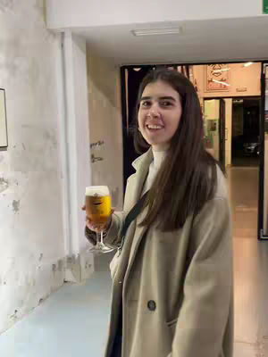 A young woman with long brown hair stands in a hallway holding a glass of beer. She is smiling and wearing a beige overcoat and a white turtleneck sweater. The background includes a door and a white wall.
