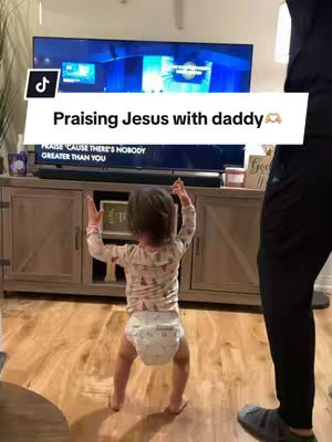 A young child stands facing the viewer with arms raised, wearing a pajama top and a diaper, in front of a television playing a video. The person filming is holding a phone in the lower right of the image. The image has hooks on the top and a tiktok logo in the top left.