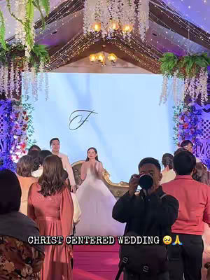 A wedding ceremony being recorded from an attendee's perspective. The foreground features people, with the wedding party in the background at the front of a well-decorated stage. The stage has a plain blue backdrop, and the attendees mostly have warm-toned clothing, with the wedding party in a white dress and suit.